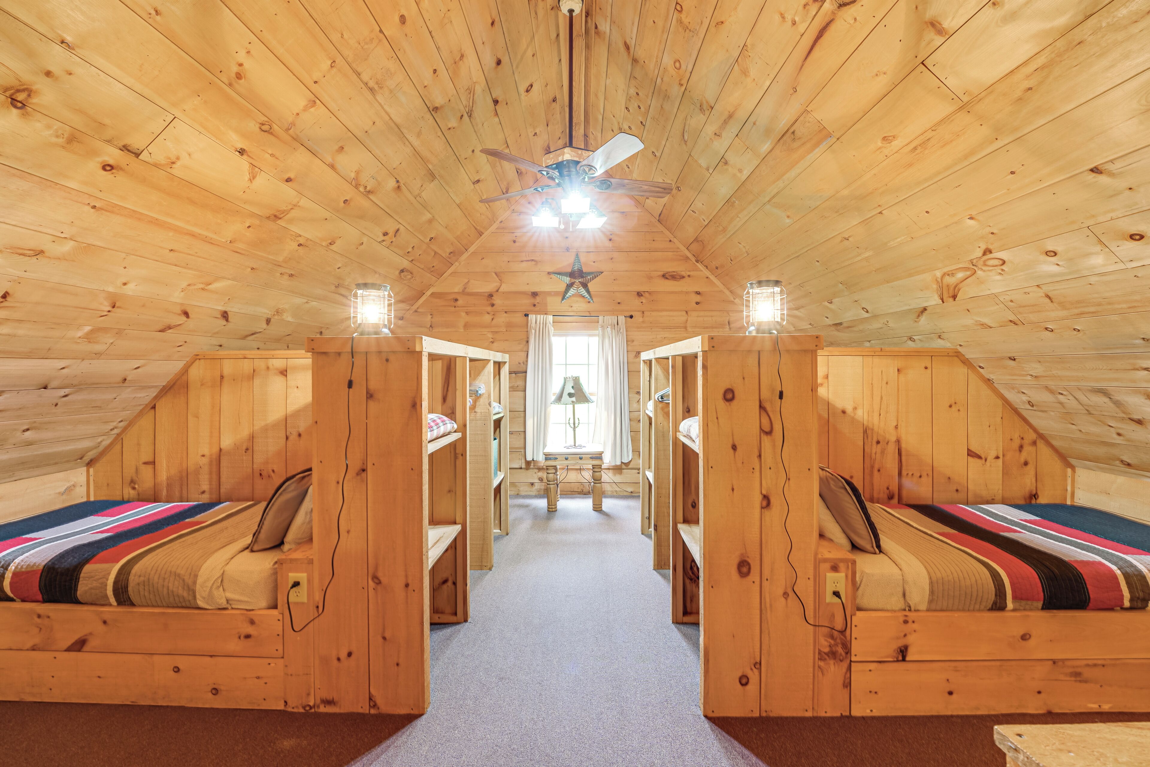 Una cabaña con techo inclinado, paredes de madera y un pasillo central que conduce a una ventana. Hay dos camas a cada lado del pasillo, cada una con una manta a rayas y almohadas. Se ve un ventilador de techo, y una decoración con forma de estrella cuelga del techo.