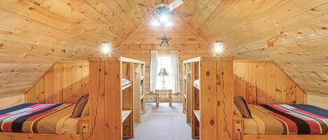 Una cabaña con techo inclinado, paredes de madera y un pasillo central que conduce a una ventana. Hay dos camas a cada lado del pasillo, cada una con una manta a rayas y almohadas. Se ve un ventilador de techo, y una decoración con forma de estrella cuelga del techo.