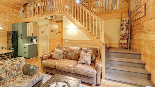 A cozy cabin interior with a wooden staircase, a leather sofa with patterned cushions, a dining table, and a kitchen area. - Fire Pit & Porch: Warm & Woodsy Rothbury Escape! (Rothbury)