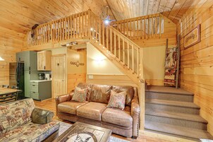 Un interior acogedor de cabaña con una escalera de madera, un sofá de cuero con cojines estampados, una mesa de comedor y una zona de cocina.