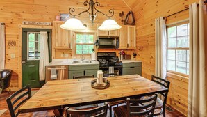 A wooden cabin interior with a dining table, chairs, a kitchen with a stove and microwave, and a sign reading "Happy Trails". - Fire Pit & Porch: Warm & Woodsy Rothbury Escape! (Rothbury)