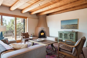 A cozy living room with a fireplace, wooden ceiling beams, and a view of the outdoors.