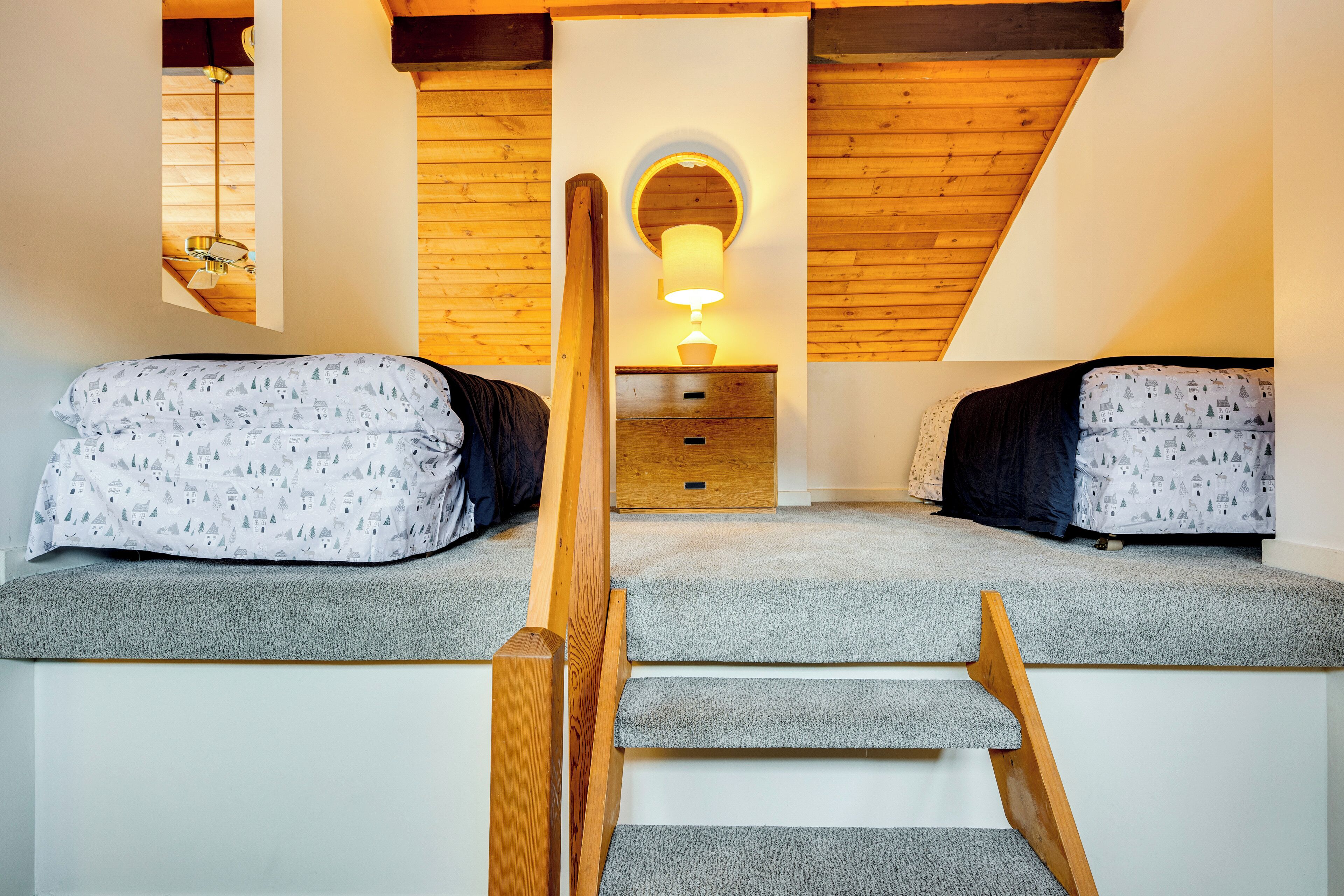 A bedroom with two beds, a wooden staircase, a small desk, and a wall-mounted lamp.