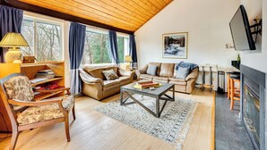 A cozy living room with wooden ceilings, a fireplace, and a large window with curtains.