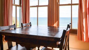 A dining table with chairs, large windows with curtains, and a view of the sea. - Beachcliff (St Austell)