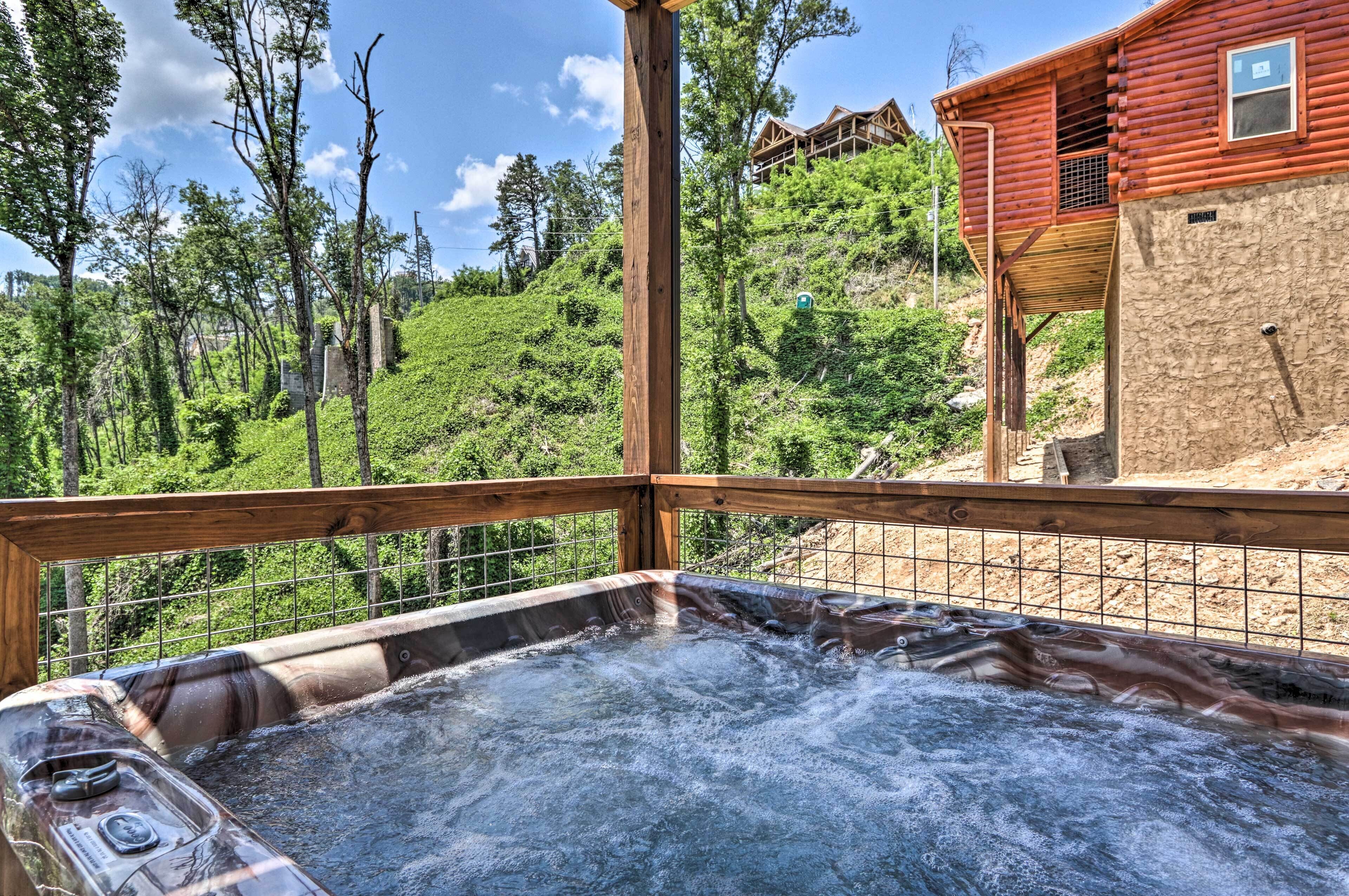 A hot tub with a wooden deck and metal railing, surrounded by lush greenery and a hillside.