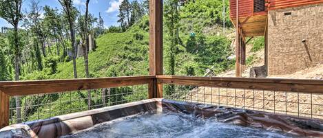 A hot tub with a wooden deck and metal railing, surrounded by lush greenery and a hillside.