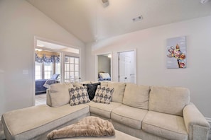 A living room with a beige sectional sofa, a fur throw, and patterned pillows. There is a door leading to another room with a bed and a window with blue curtains. A painting is on the wall. - 1 Mi to Myrtle Beach Access: Condo w/ Resort Perks (Myrtle Beach)