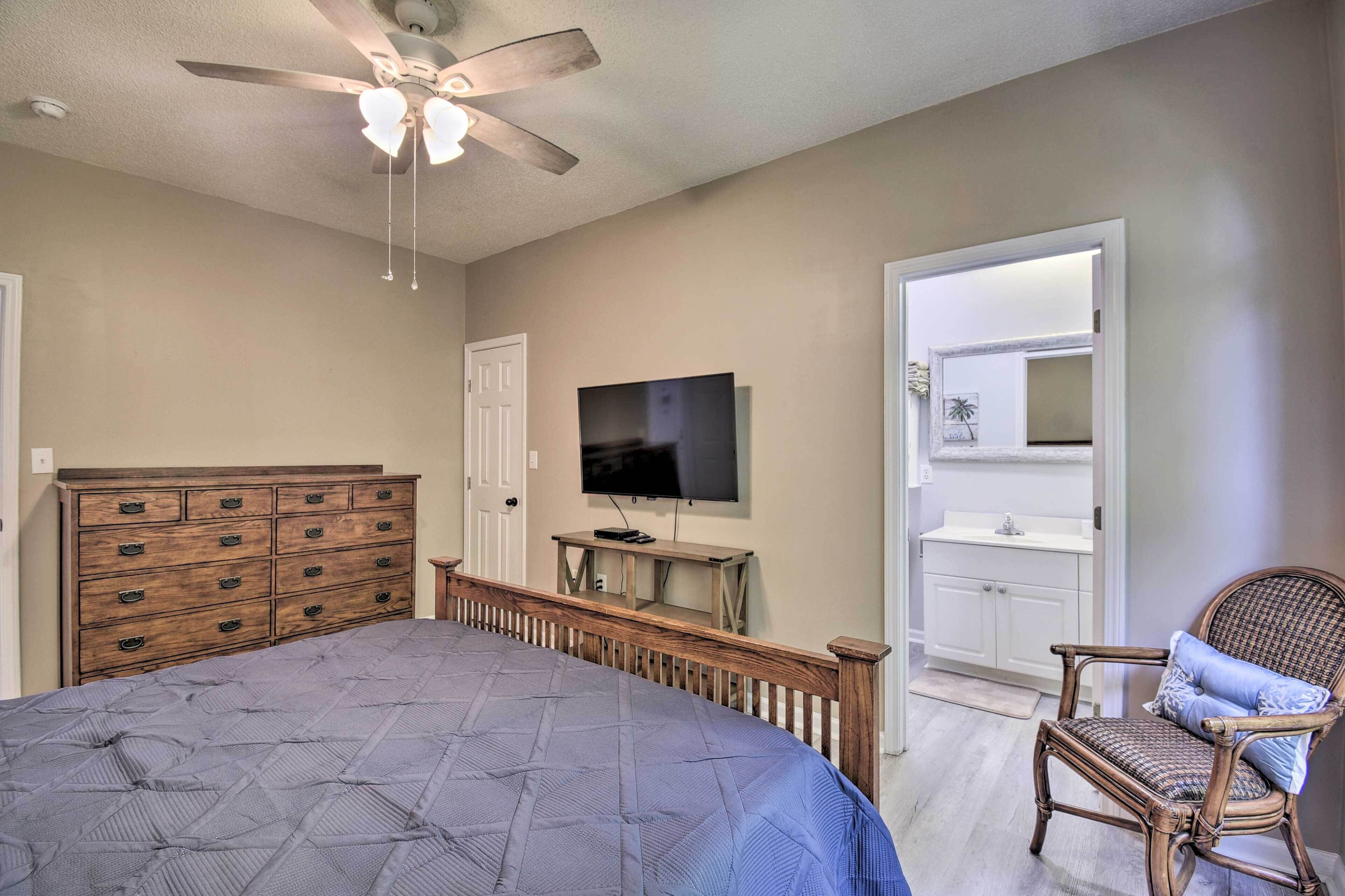 A bedroom with a bed, dresser, TV, chair, and a door leading to a bathroom.