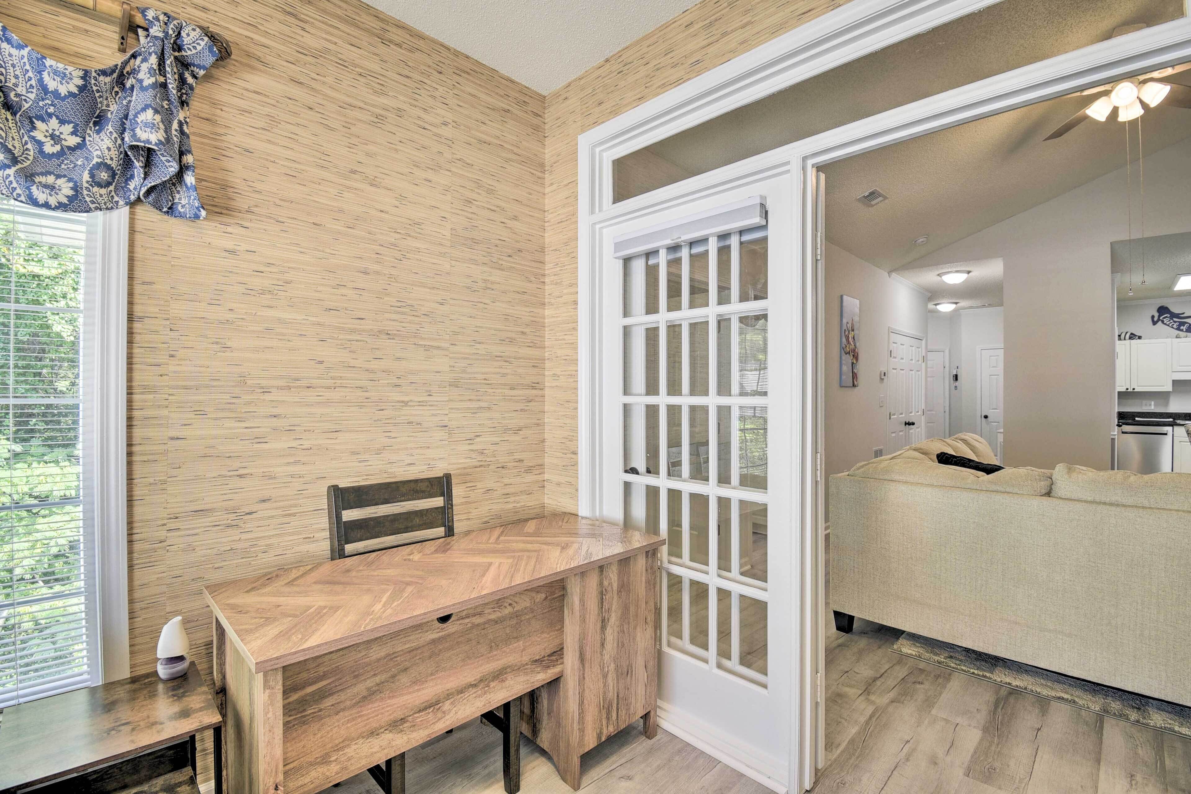 A room with a wooden herringbone floor, a wooden desk, a chair, a sofa, and a large window with blinds.