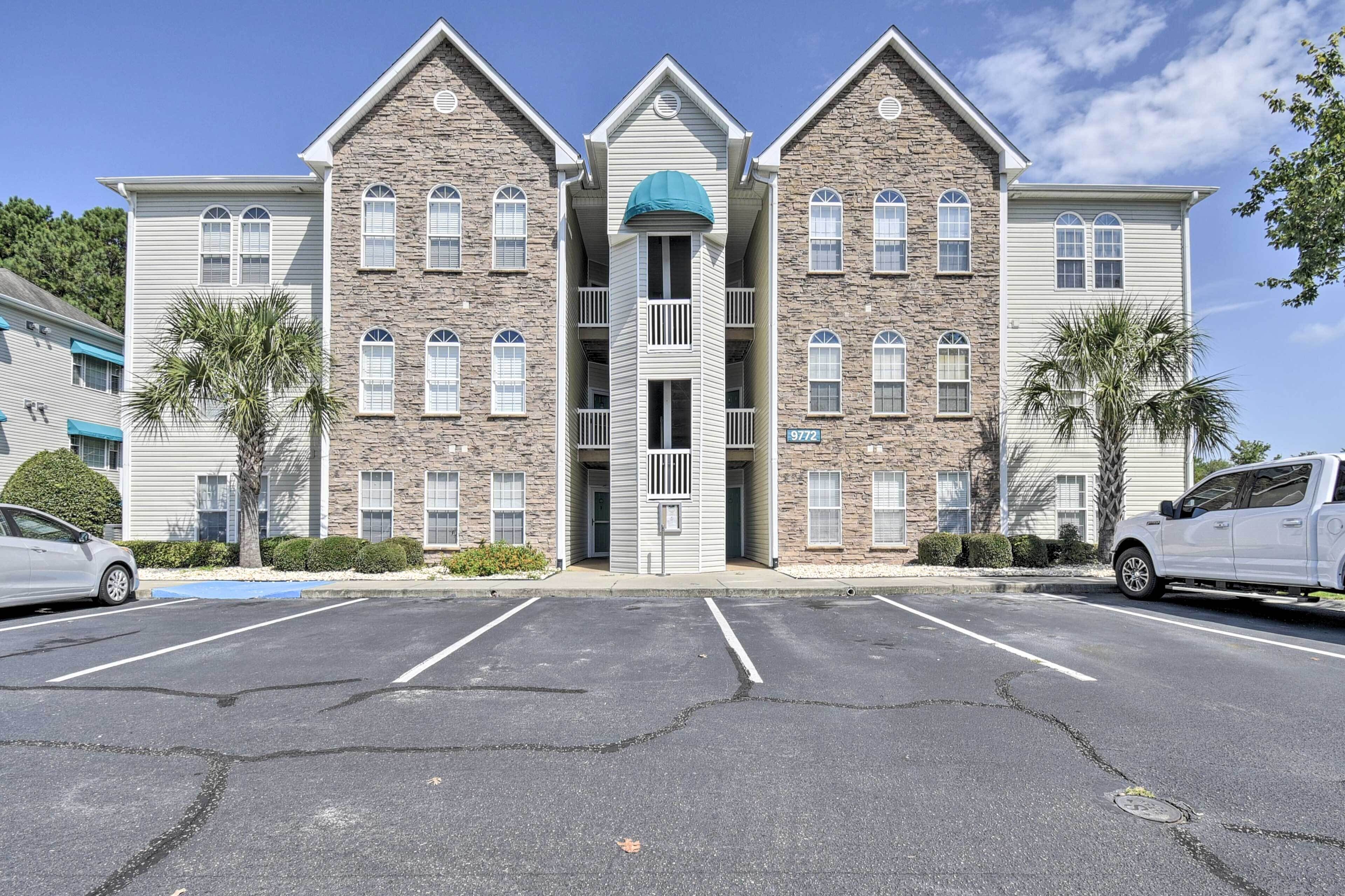 A multi-story building with a central entrance, parking lot, and two cars parked in front.