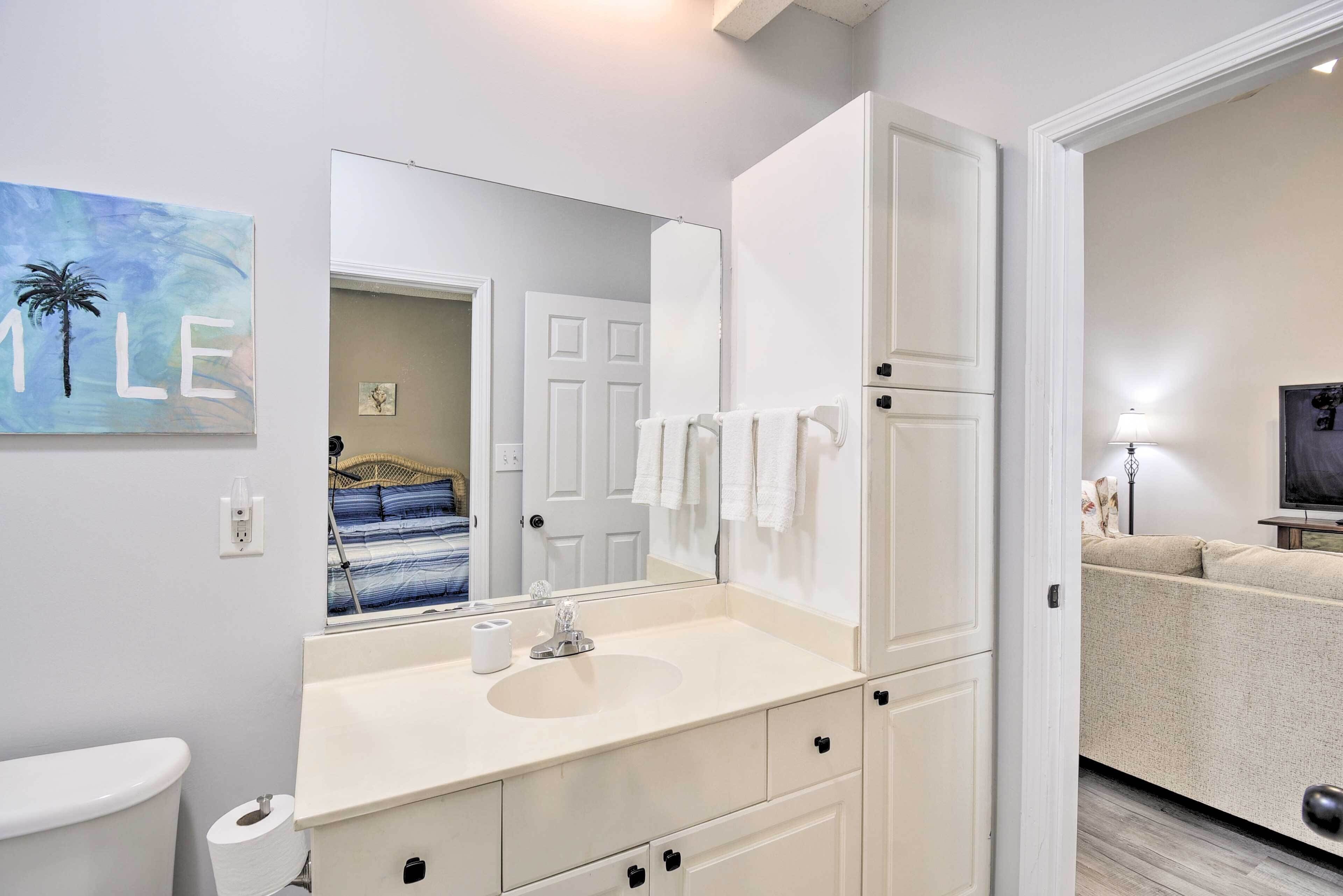 A modern bathroom with a white sink, mirror, and cabinets. There is a toilet to the left and a door leading to another room.