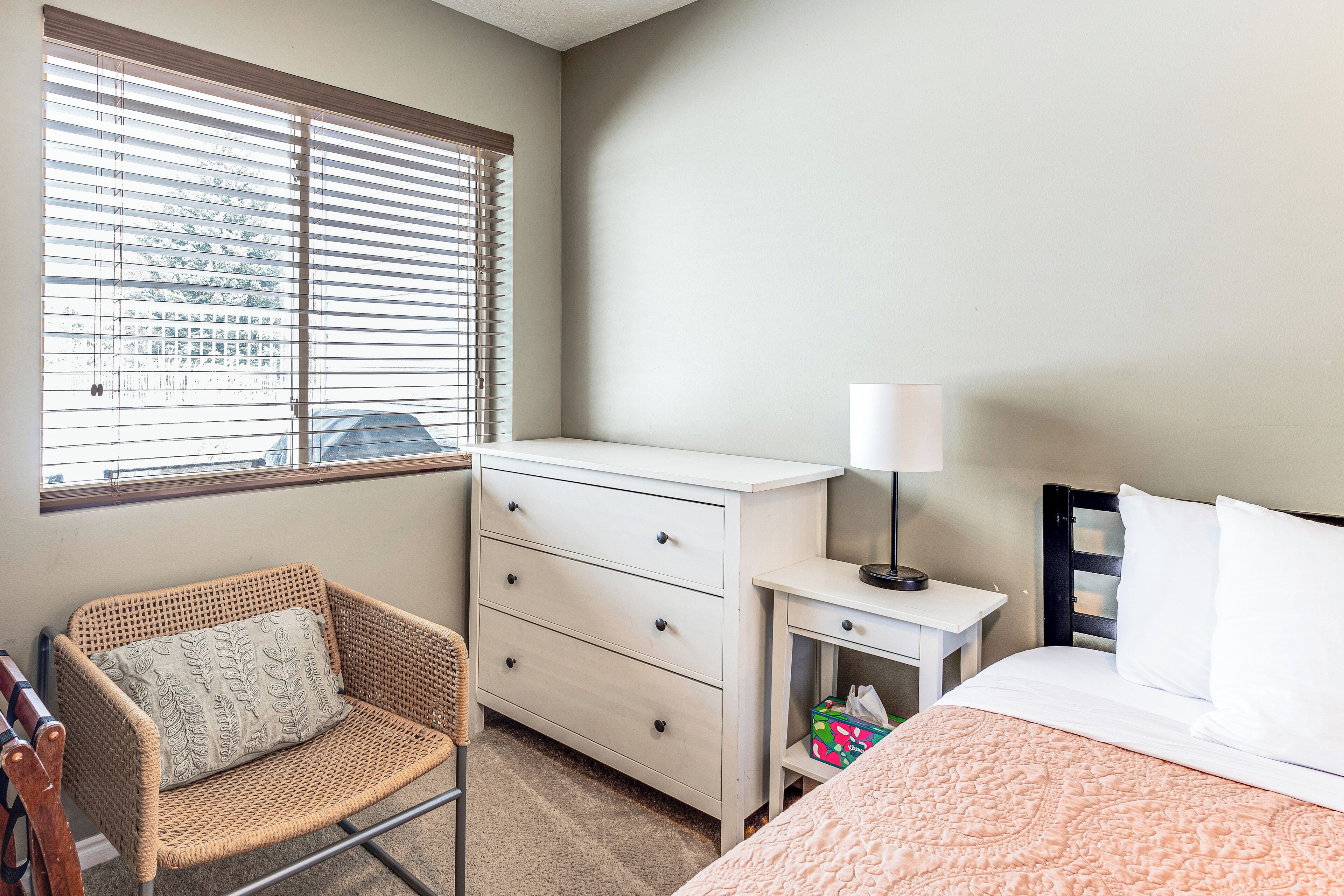 A bedroom with a bed, a dresser, a chair, a nightstand, and a window with blinds.