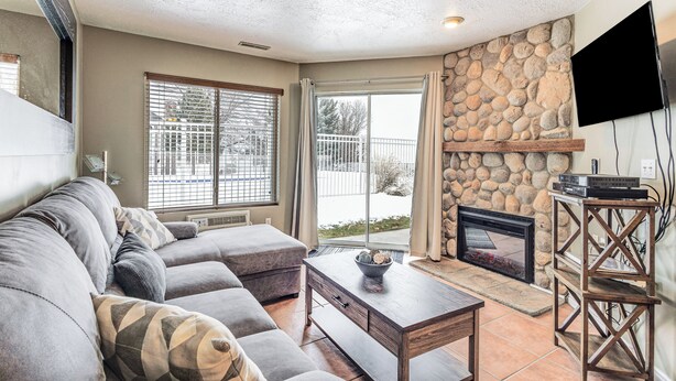 A living room with a stone fireplace, a gray sectional sofa, and a flat-screen TV mounted on the wall.