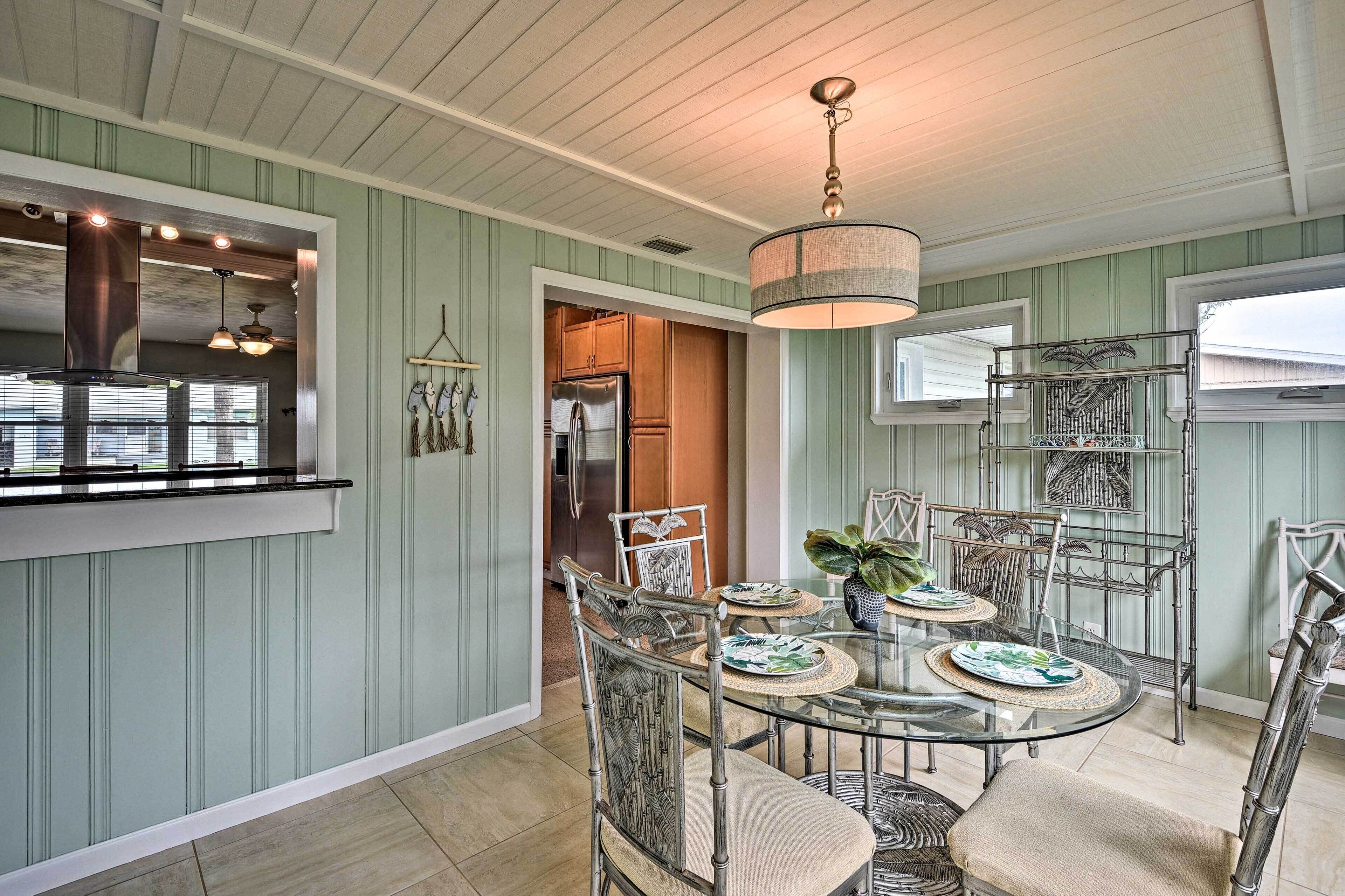 A dining area with a glass table and metal chairs, a hanging light fixture, and a kitchen in the background.