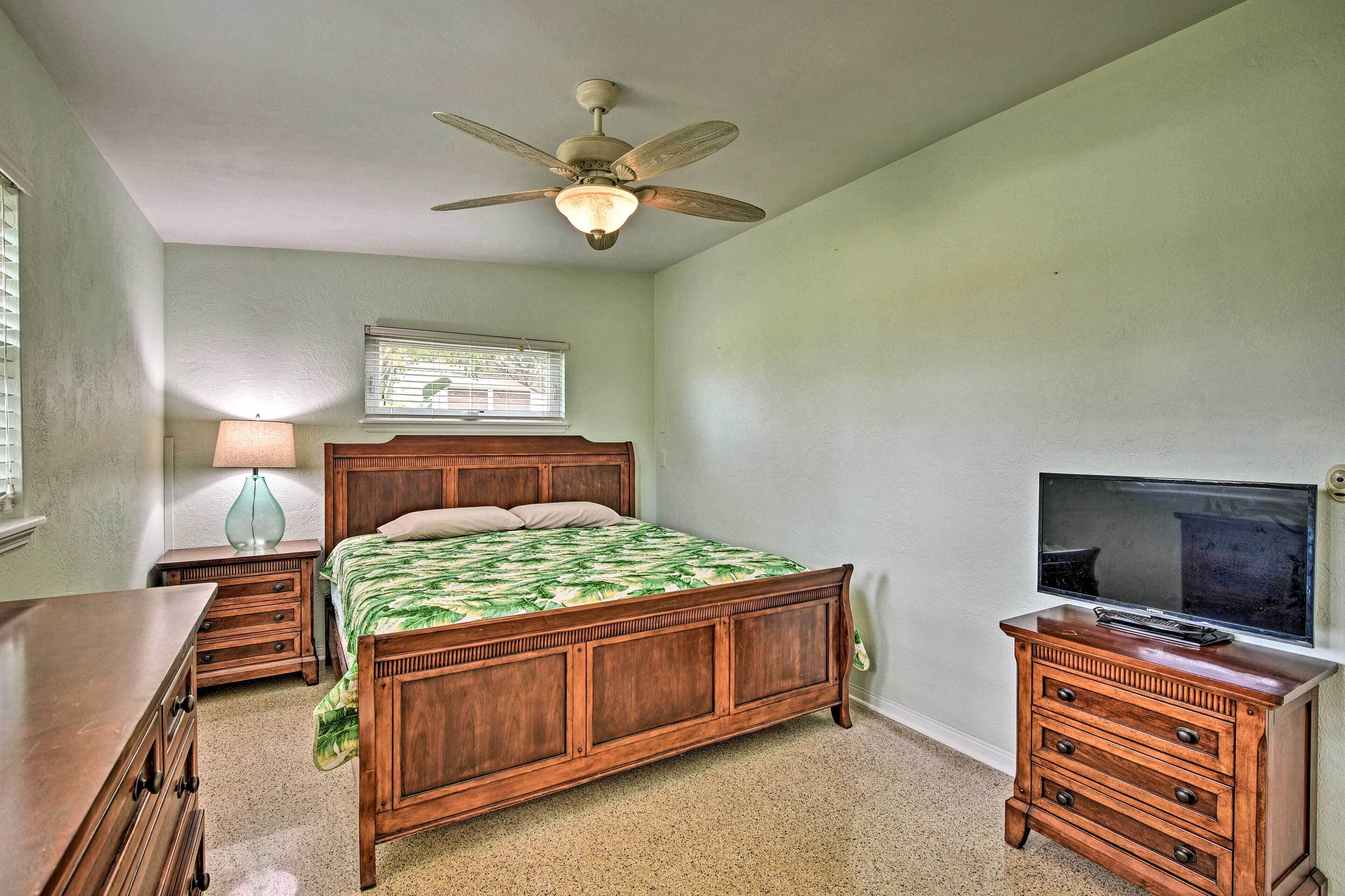 A bedroom with a wooden bed, a ceiling fan, a TV on a dresser, and a window with blinds.