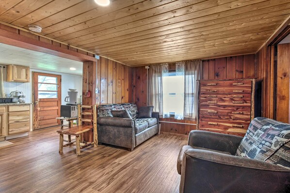 A wooden cabin interior with a sofa, a chair, a wooden cabinet, and a kitchen area. - Lakefront Minocqua Cabin w/ Dock, Wifi & AC (Minocqua)