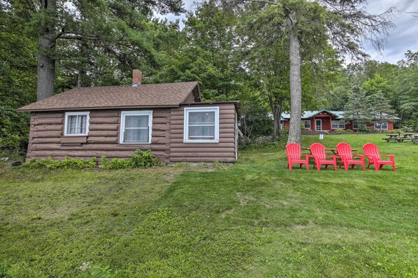 A log cabin with two windows and a chimney, surrounded by a grassy area with red chairs and a picnic table. - Lakefront Minocqua Cabin w/ Dock, Wifi & AC (Minocqua)