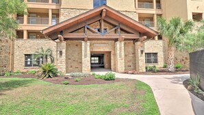 A multi-story building with a stone facade, a central entrance with a wooden overhang, and a landscaped courtyard with grass and small plants. - Lake LBJ Condo w/ Balcony & Shared Boat Docks (Horseshoe Bay)