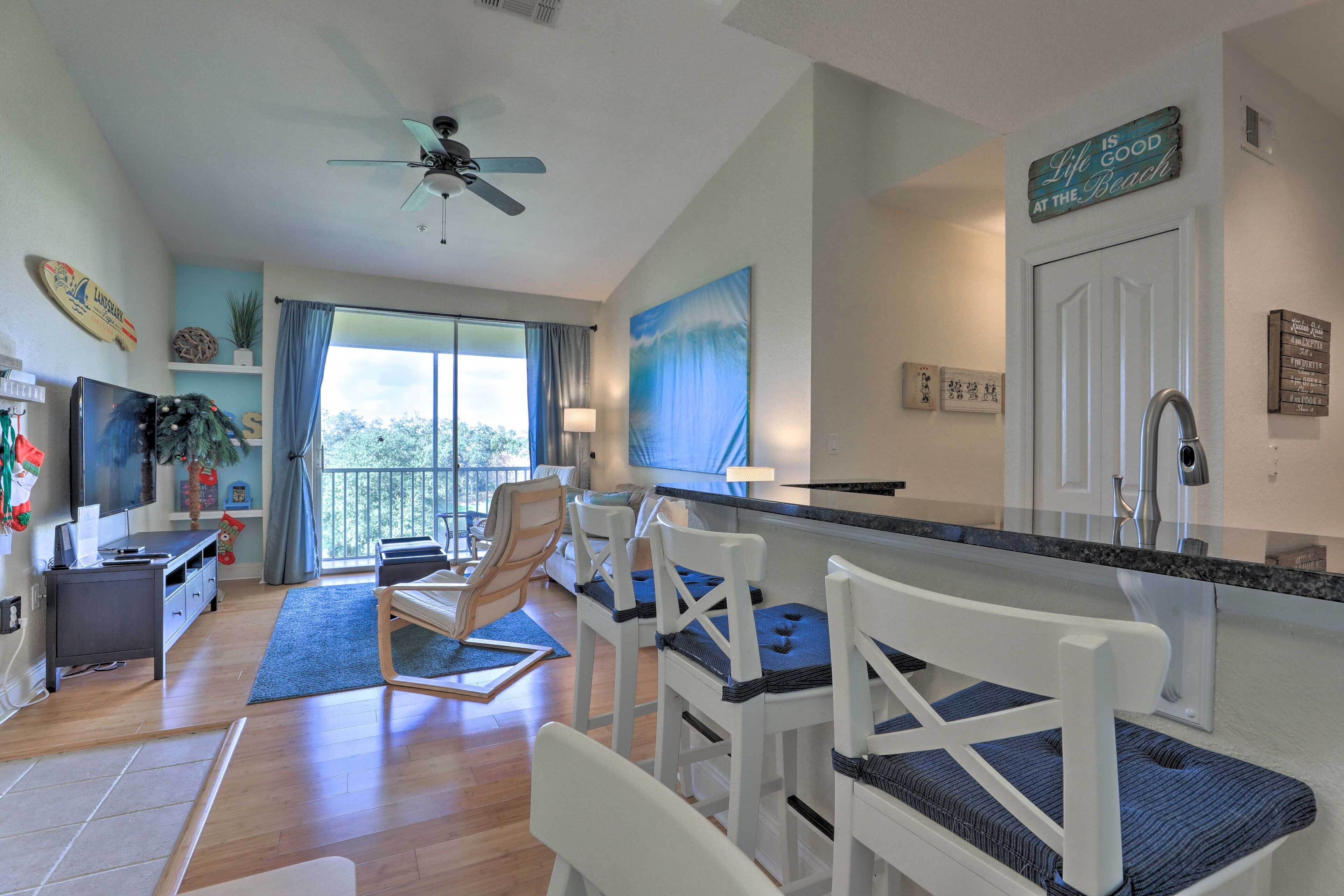 A spacious living area with a large window, a ceiling fan, and a kitchen counter with bar stools.