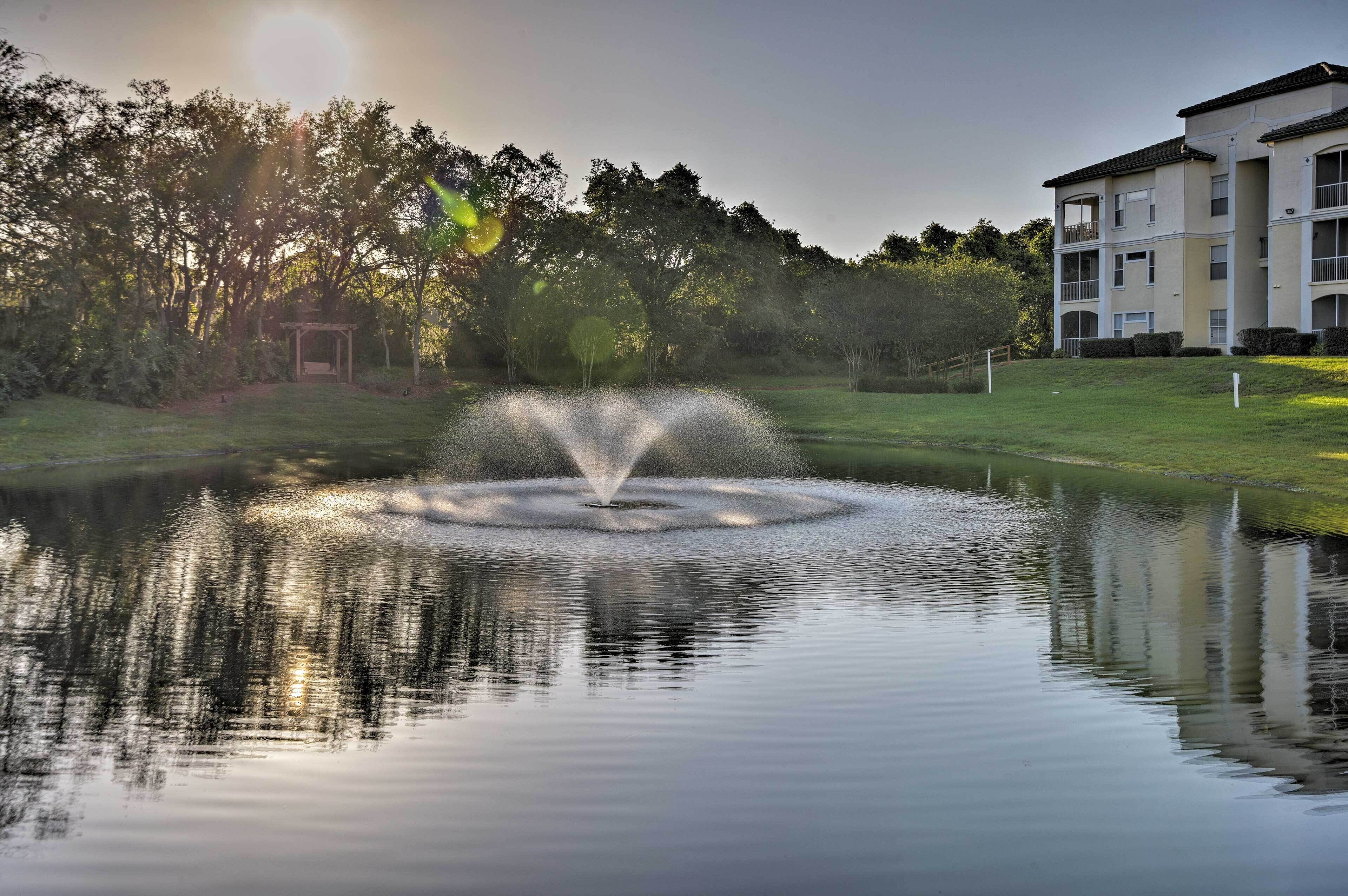 A tranquil pond with a fountain, surrounded by a well-maintained lawn and trees. A multi-story building is visible in the background.
