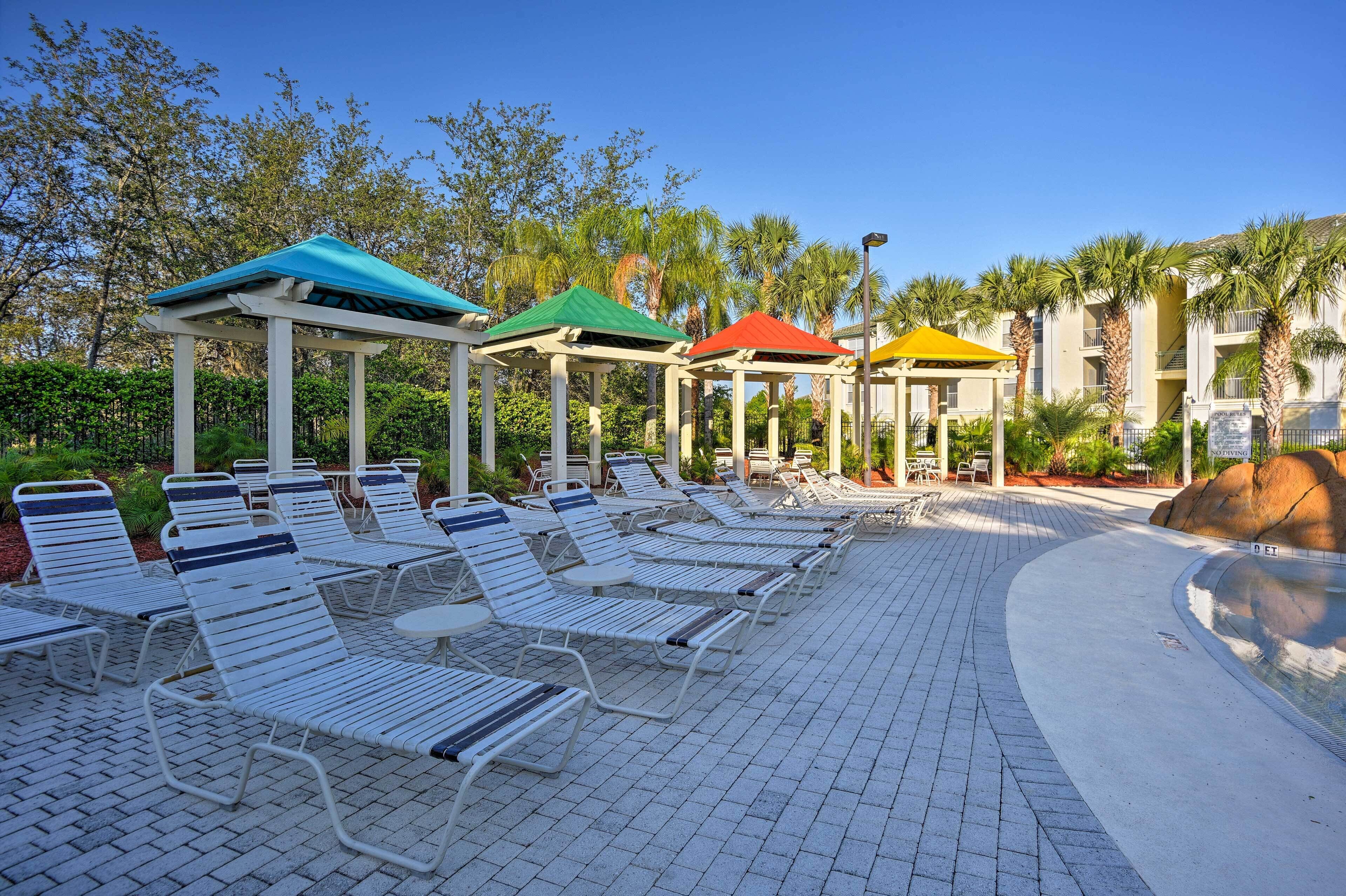 Un espace aménagé au bord de la piscine, avec des chaises longues blanches et des parasols colorés, entouré de palmiers et surplombé d’un ciel dégagé.