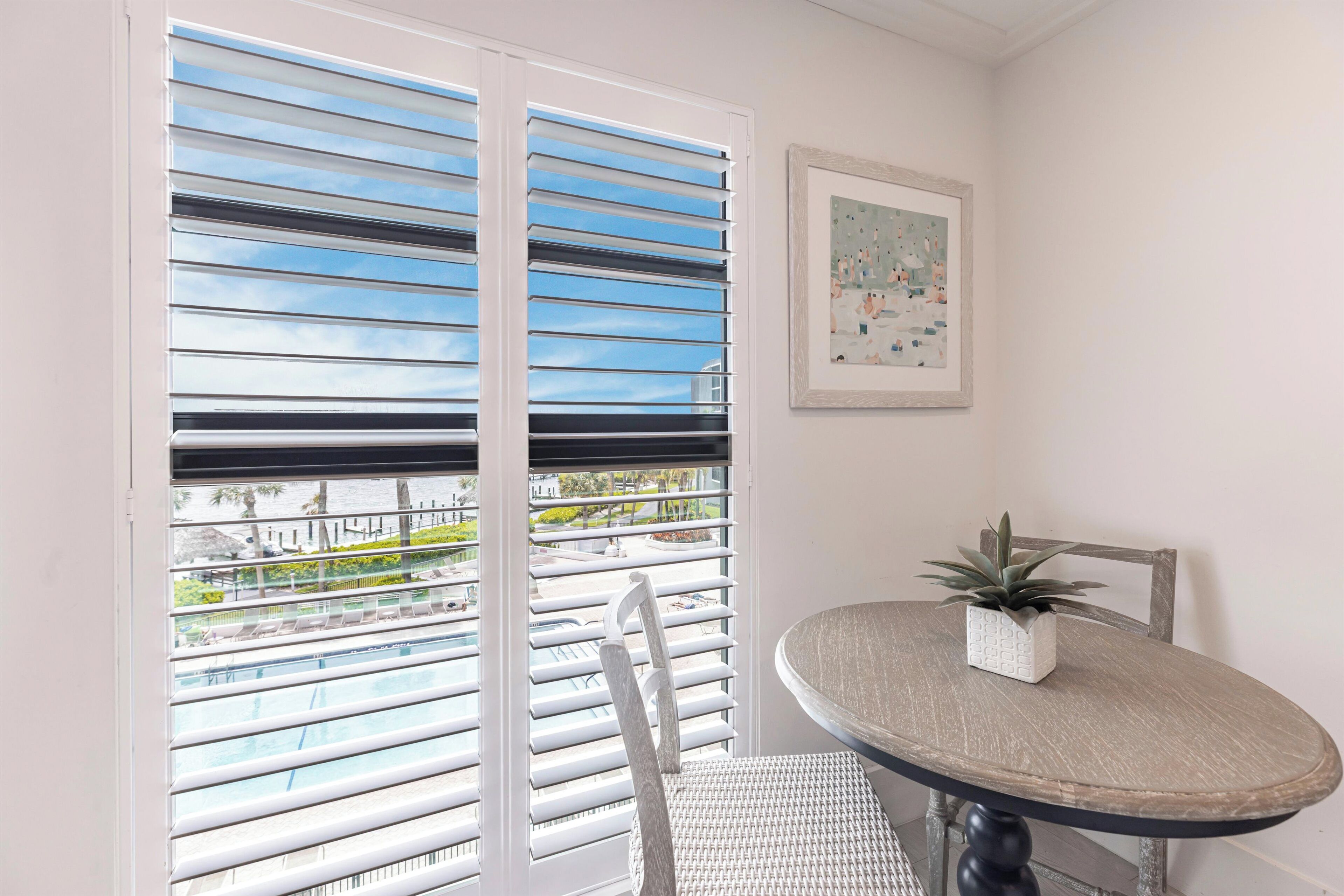 A balcony with white blinds, a wicker chair, a round table with a potted plant, and a framed picture on the wall.