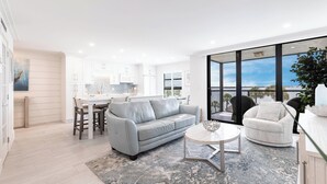 Een moderne woonkamer met een grijze bank, een ronde salontafel en een witte fauteuil. Achtergrond toont een keuken met witte kasten en een eethoek met een tafel en stoelen.