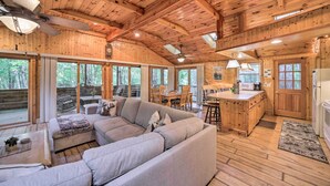 L’intérieur d’une cabane en rondins, avec un grand canapé d’angle gris, un plancher en bois, un coin cuisine et de grandes fenêtres.