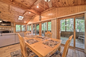 A wooden cabin interior with a dining table, chairs, a sofa, and a stone fireplace. - Fire Pit & Grill: Cozy Cabin in Lake Toxaway! (Lake Toxaway)