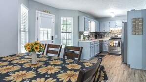 A dining area with a table set for two, a vase of flowers, and a kitchen with light blue cabinets and a central island. - Above-ground Pool & Backyard at Murfreesboro Home! (Murfreesboro)