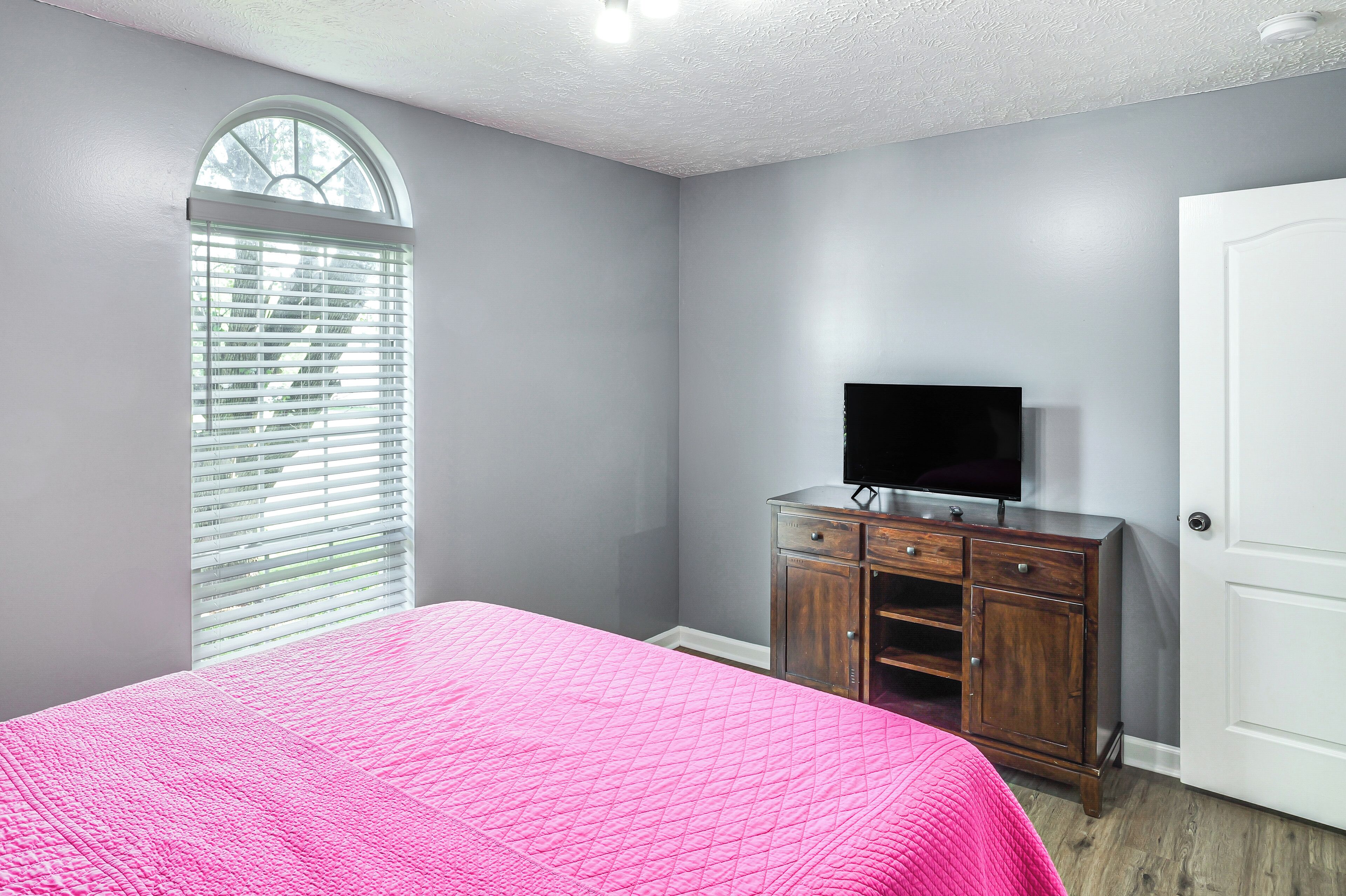 Une chambre avec une couvre-lit rose matelassé, une commode en bois surmontée d’un téléviseur à écran plat, une porte blanche et une fenêtre avec des stores.
