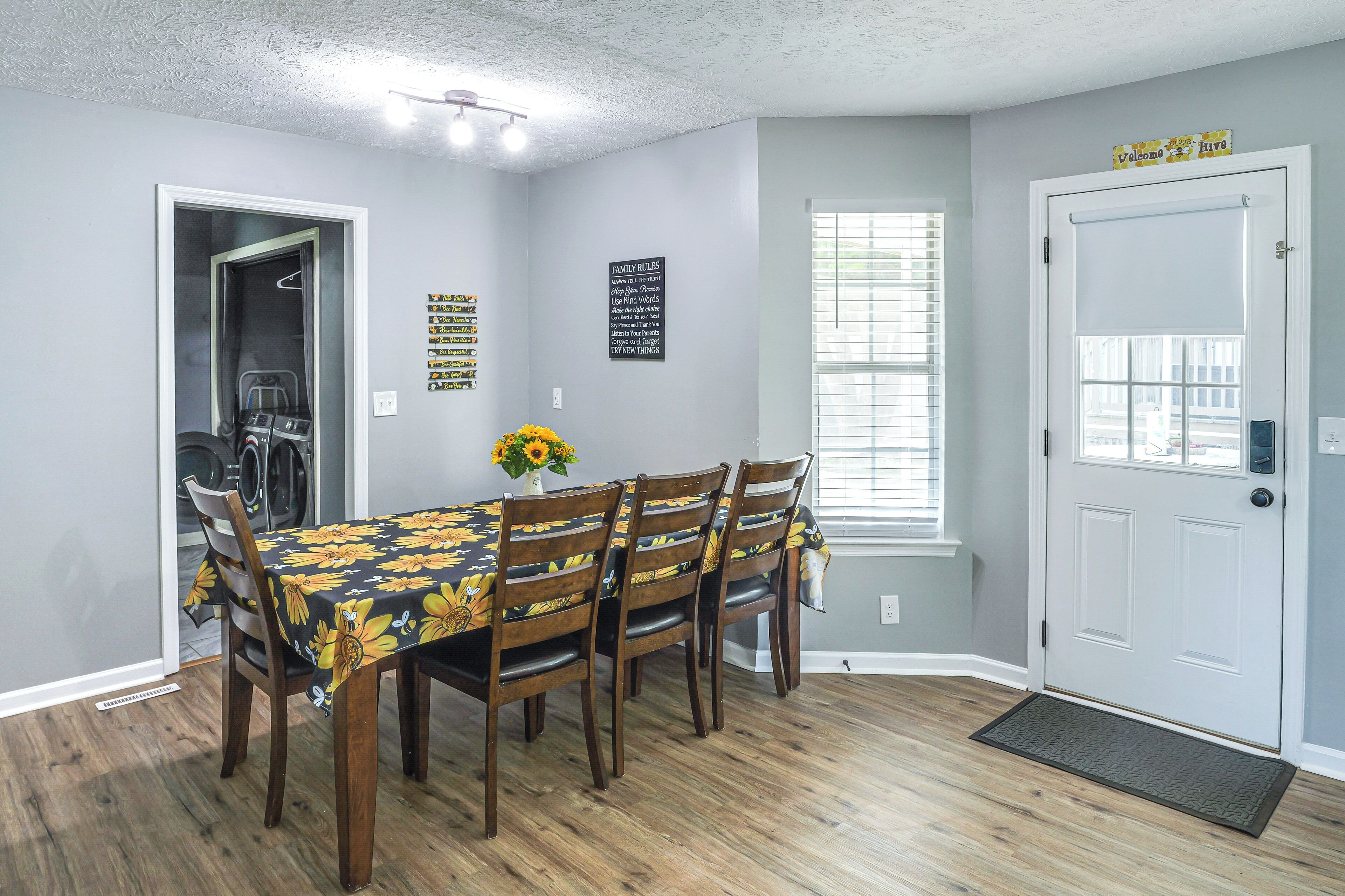 Un comedor con una mesa dispuesta para cuatro personas, un jarrón con girasoles y una puerta con un letrero que dice 'Welcome'.