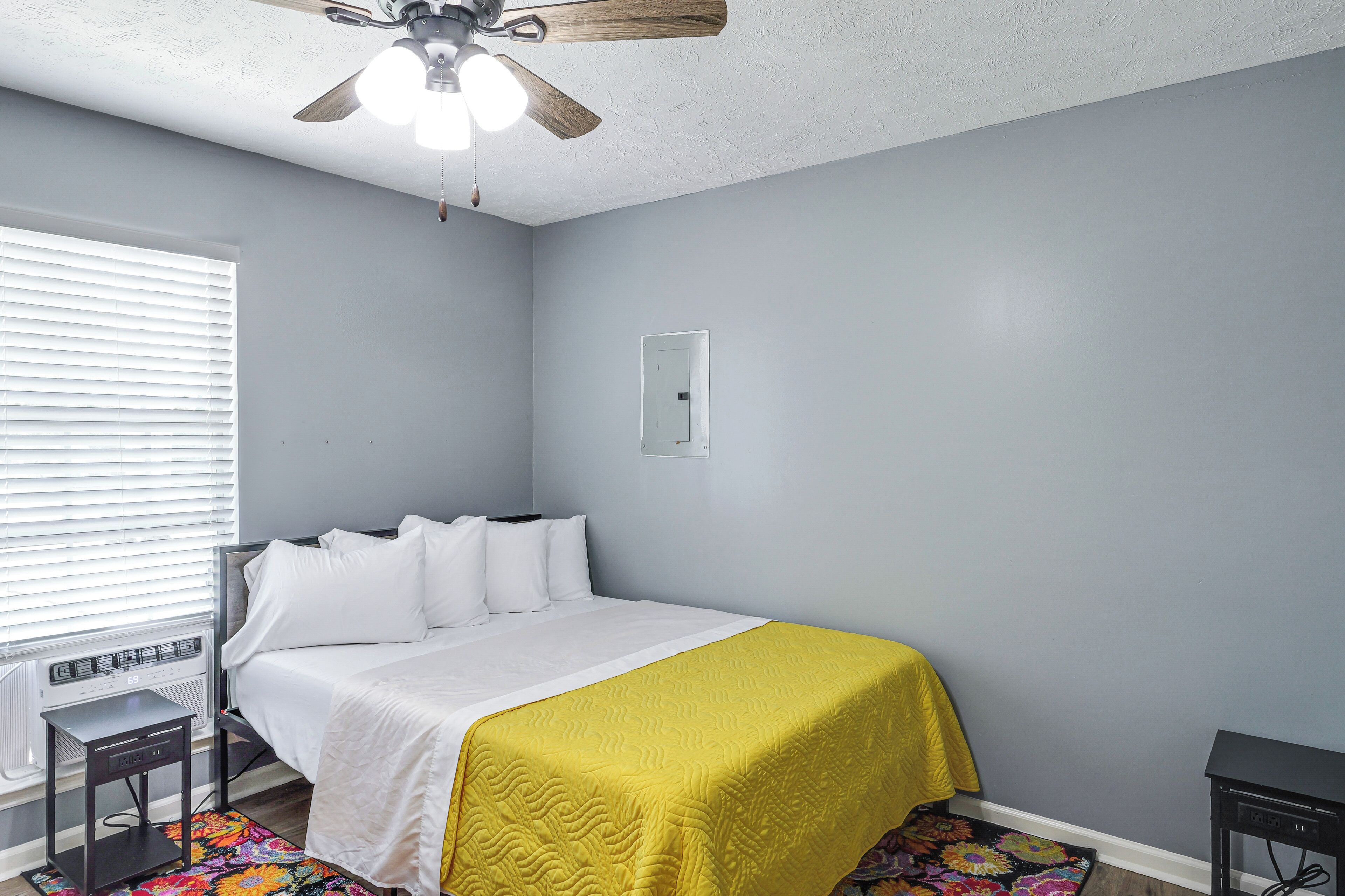 Une chambre à coucher avec un lit, un ventilateur de plafond, une fenêtre avec des stores et une petite table.