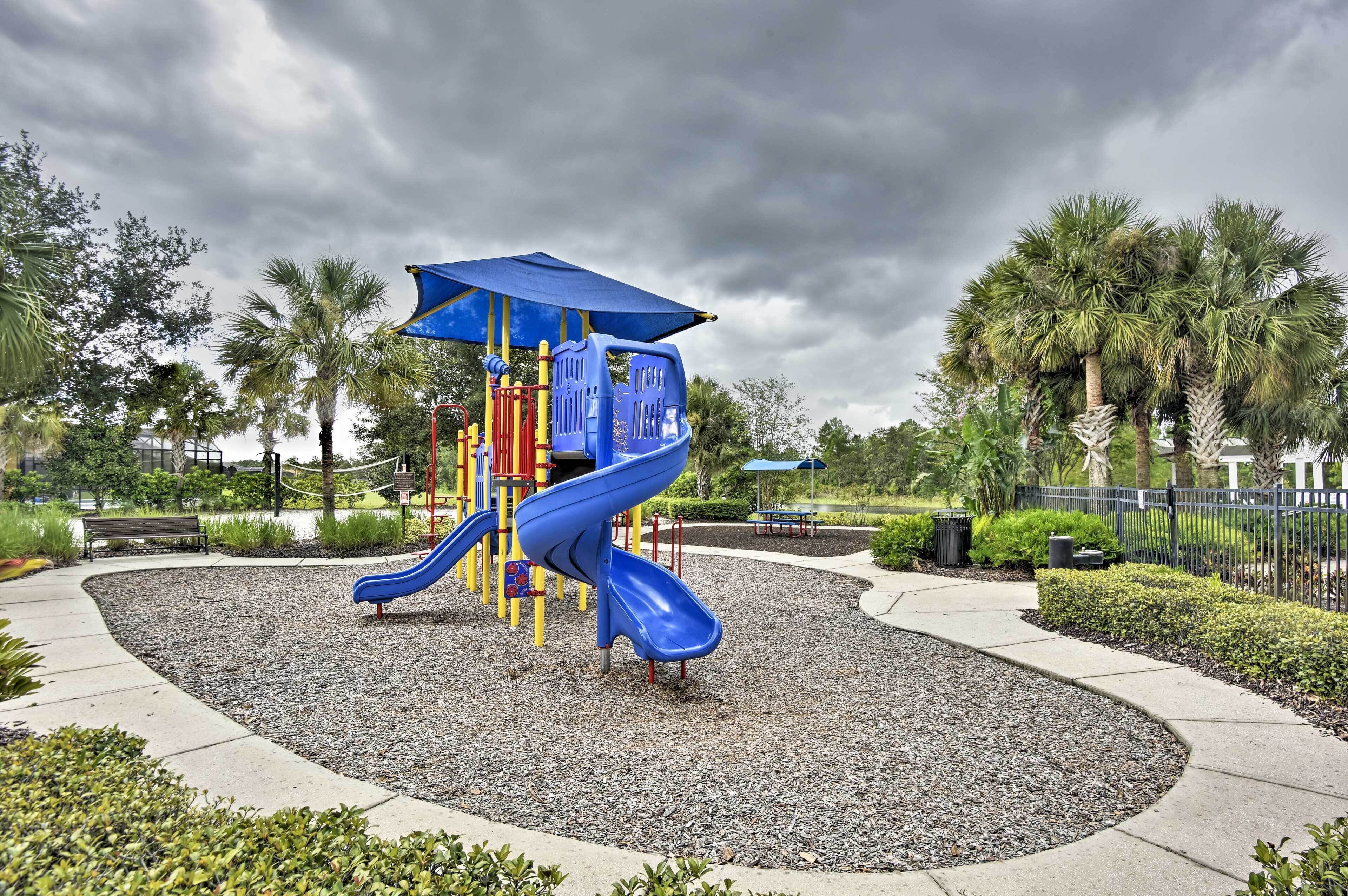A playground with a blue slide and climbing structures, surrounded by gravel and palm trees.