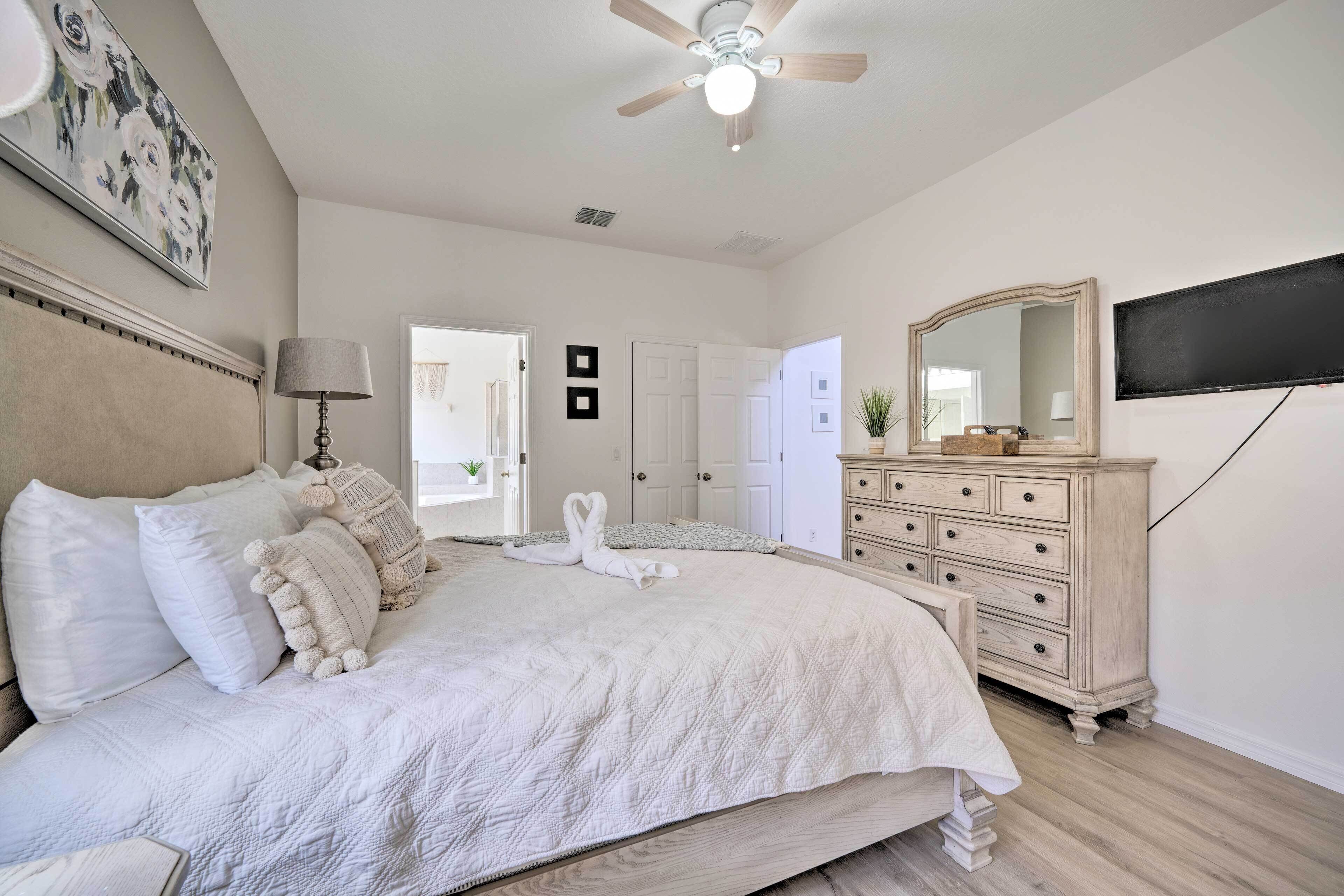 Une chambre avec un grand lit, un ventilateur de plafond, une commode et un téléviseur fixé au mur.
