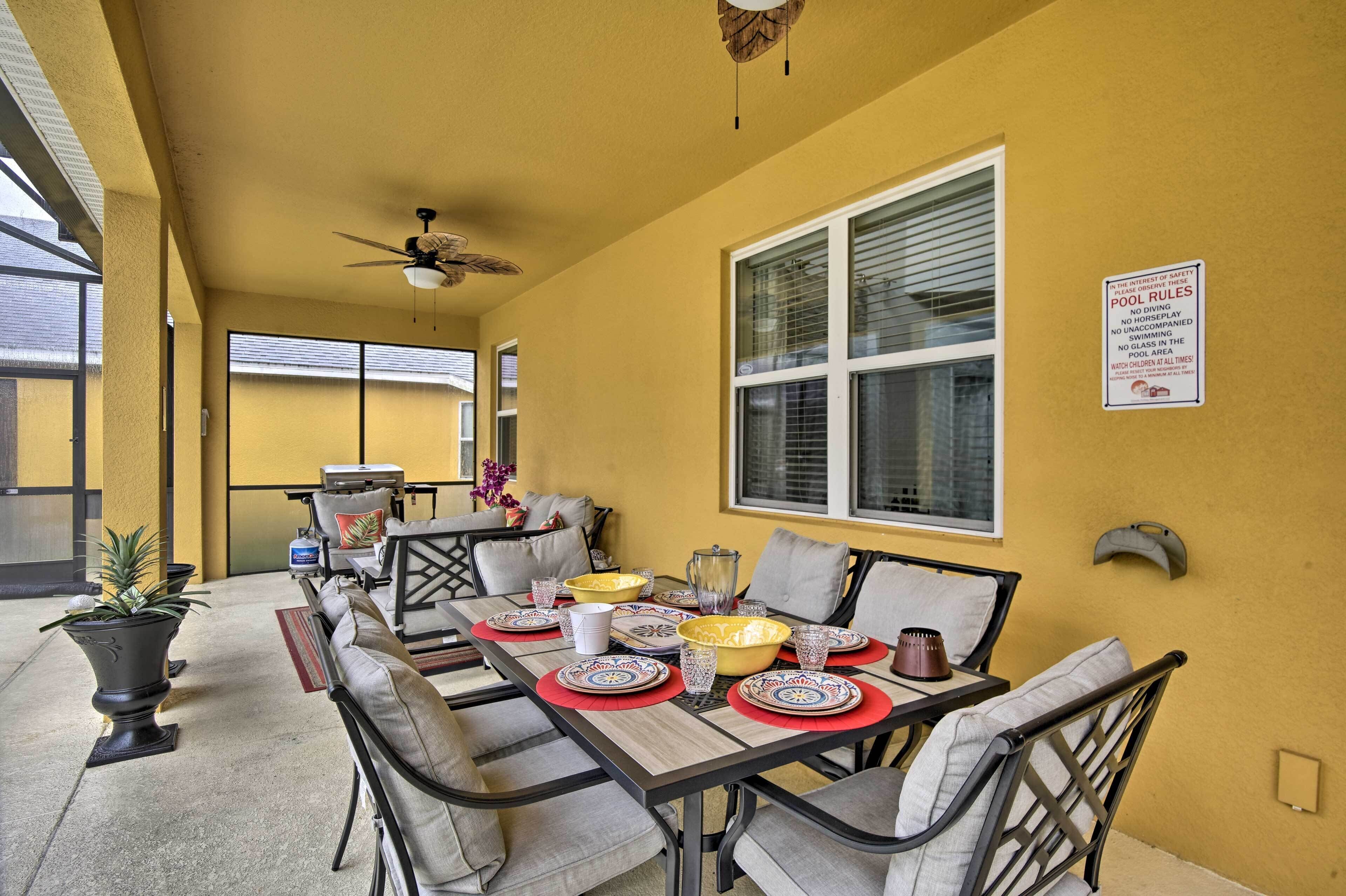 Un coin repas avec une table dressée pour six personnes, des chaises, un ventilateur de plafond et une fenêtre avec des stores.