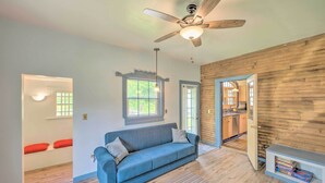 A modern living room with a blue sofa, wooden walls, a ceiling fan, and a view into the kitchen. - Rustic & Historic Guildhall Farmhouse w/ Fire Pit! (Guildhall)