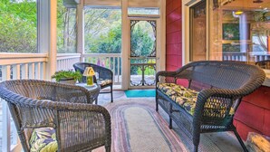 A covered porch with wicker furniture, a view of trees, and a red wall. - Mtn Views + Fire Pit: Serene Chimney Rock Abode (Chimney Rock)