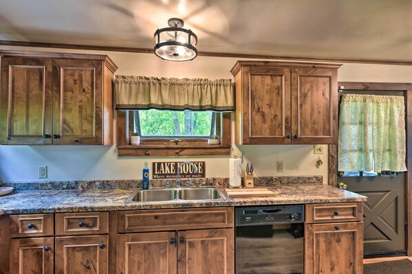 A kitchen with wooden cabinets, a granite countertop, a dishwasher, and a window with a view of trees.