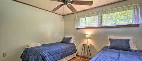 Un dormitorio con dos camas, un ventilador de techo, una ventana con cortinas de encaje y una mesita de noche pequeña con una lámpara.