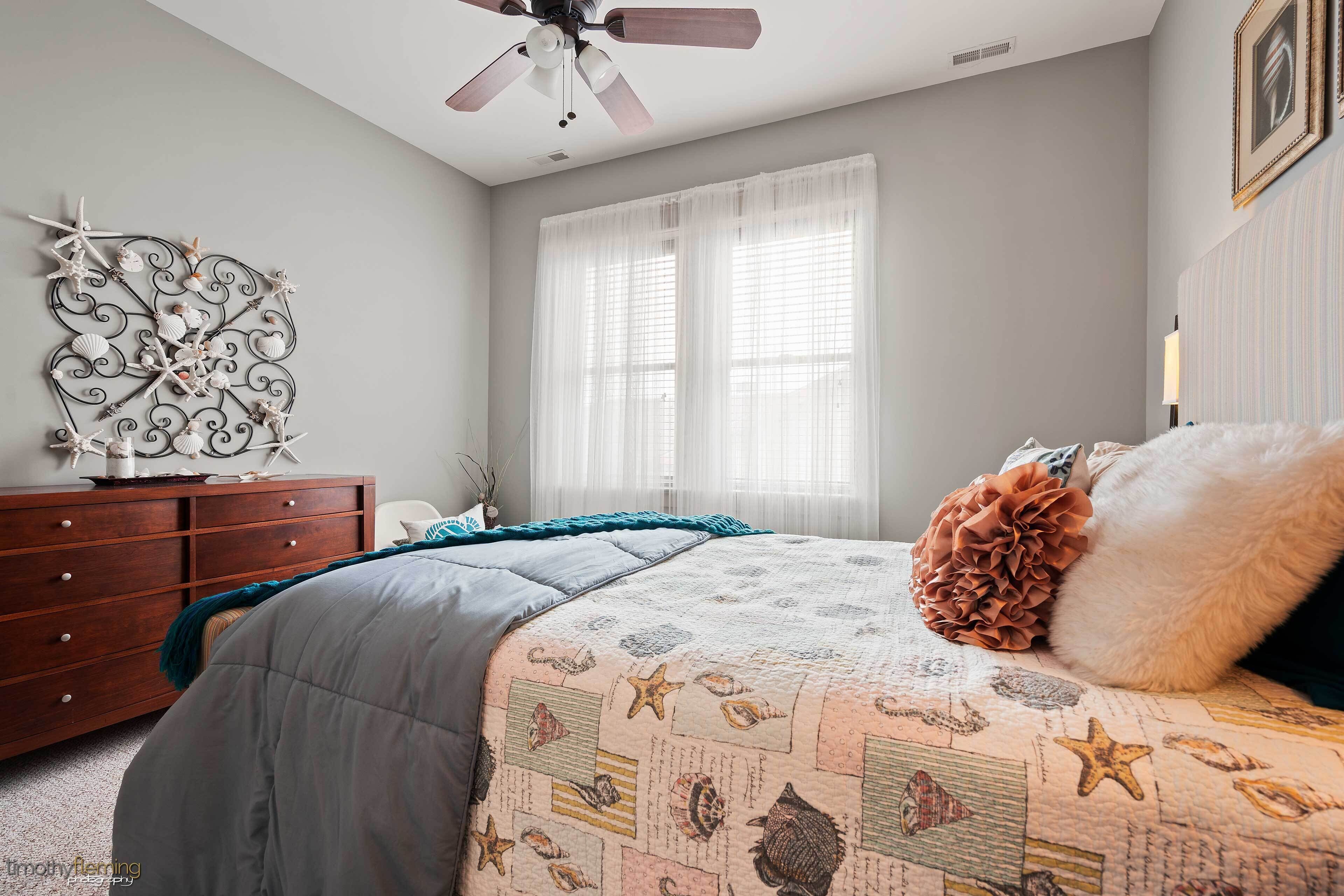 A bedroom with a bed, a dresser, a ceiling fan, and a window with sheer curtains.