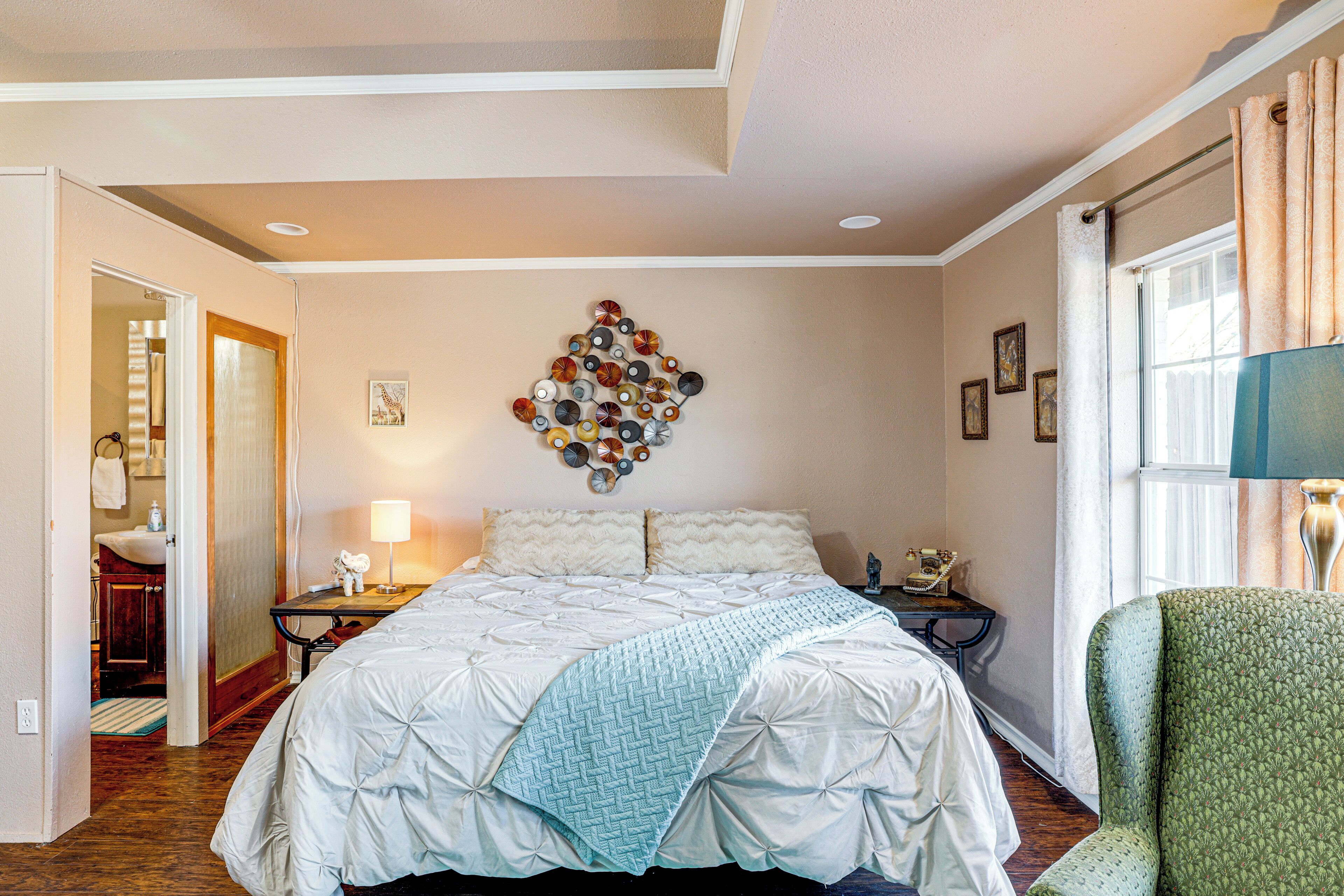 A bedroom with a bed, a nightstand, a lamp, a chair, a wall art piece, and a window with curtains.