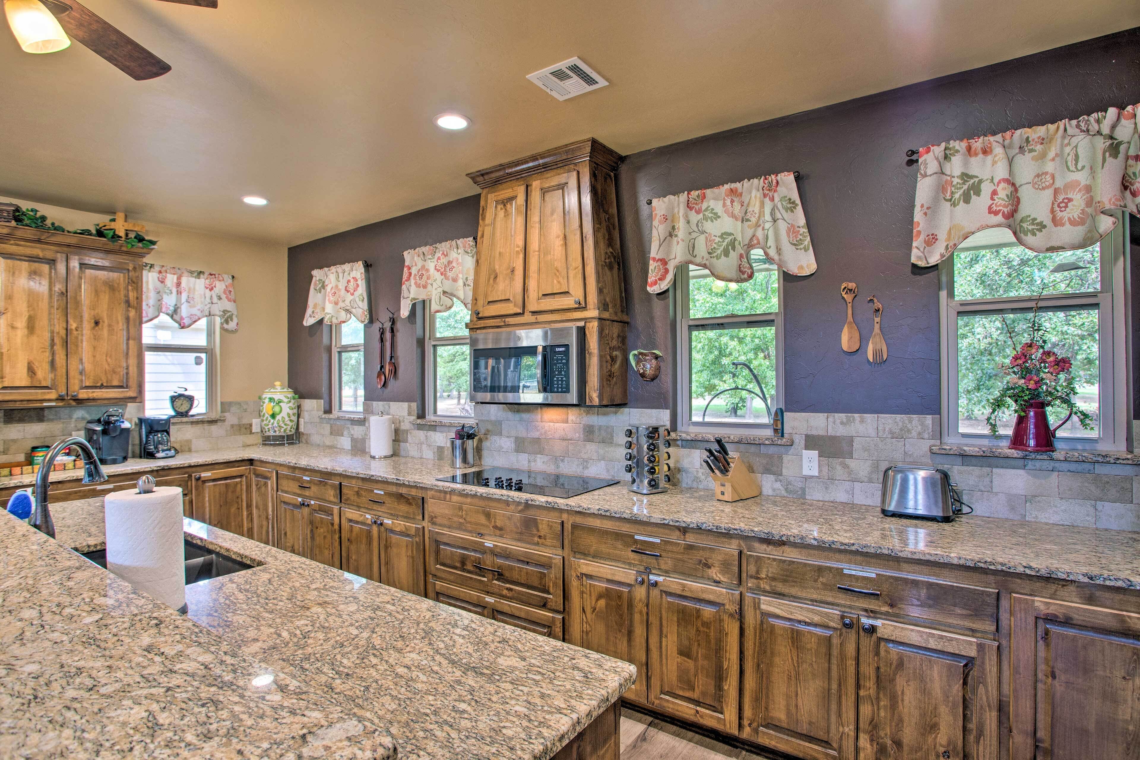 A kitchen with granite countertops, wooden cabinets, a stainless steel microwave, and a window with a view of greenery.