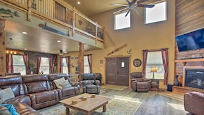 A spacious living room with a large sectional sofa, a wooden coffee table, and a fireplace. There is a balcony with a railing and a ceiling fan. - Kingston Home w/ Pickleball: Steps to Lake Texoma! (Kingston)