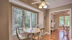 A dining area with a round table and four chairs, large windows with a view of trees, a ceiling fan, and a kitchen area in the background. - Lakefront Getaway w/ Private Dock & Kayaks! (Bella Vista)