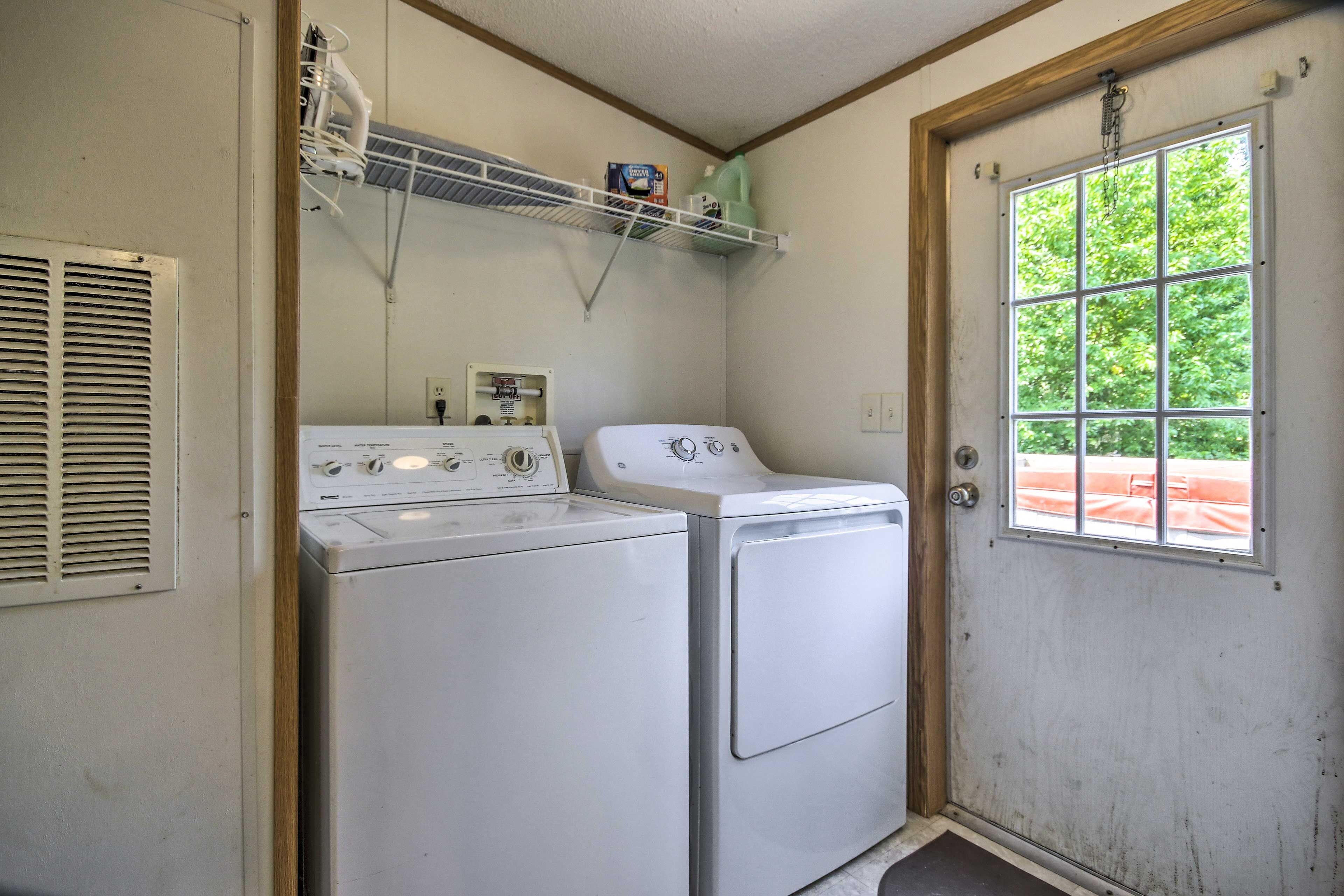 A laundry room with two white washers and dryers, a vented dryer, a shelf with cleaning supplies, and a window with a view of greenery.