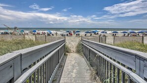 En træpromenade der fører til en strand med blå parasoller og en sandstrand.