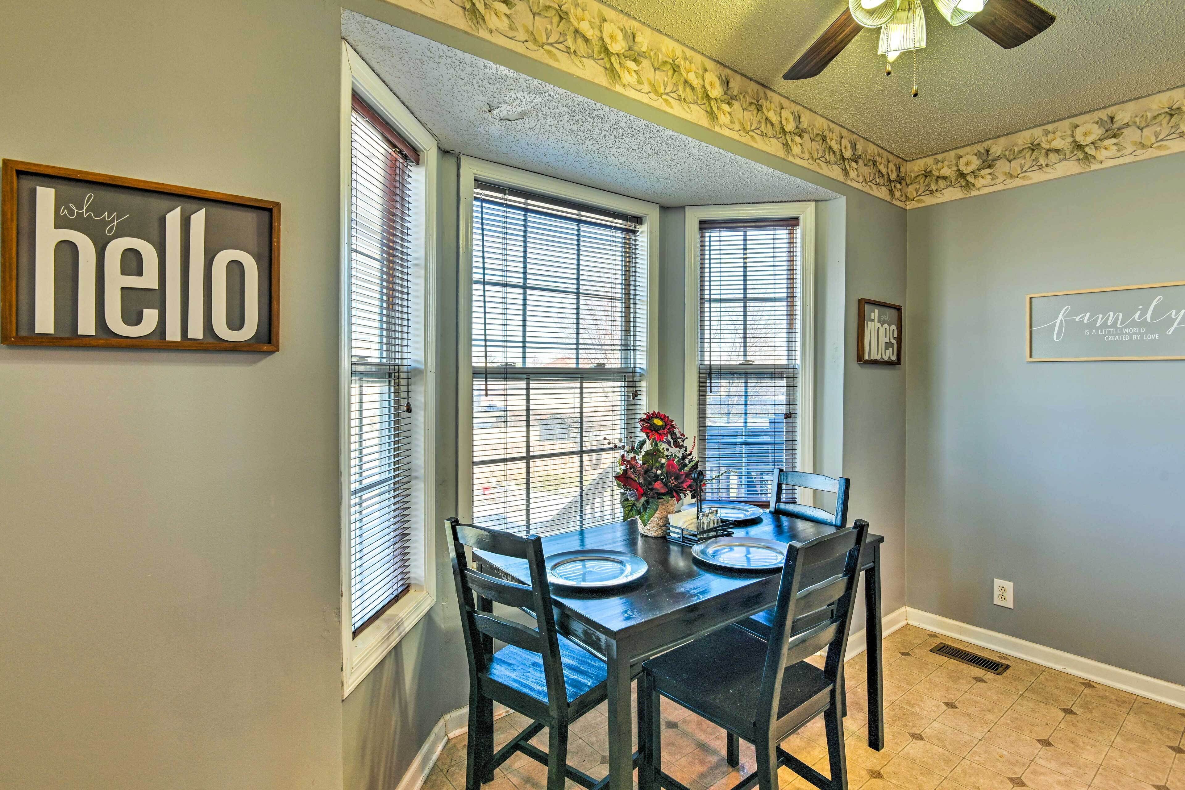 A dining area with a table set for four, flanked by three windows with blinds, a ceiling fan, and a wall with decorative floral wallpaper.