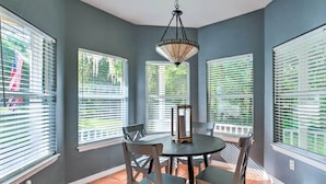 A dining area with a round table and four chairs, large windows with blinds, and a hanging light fixture.